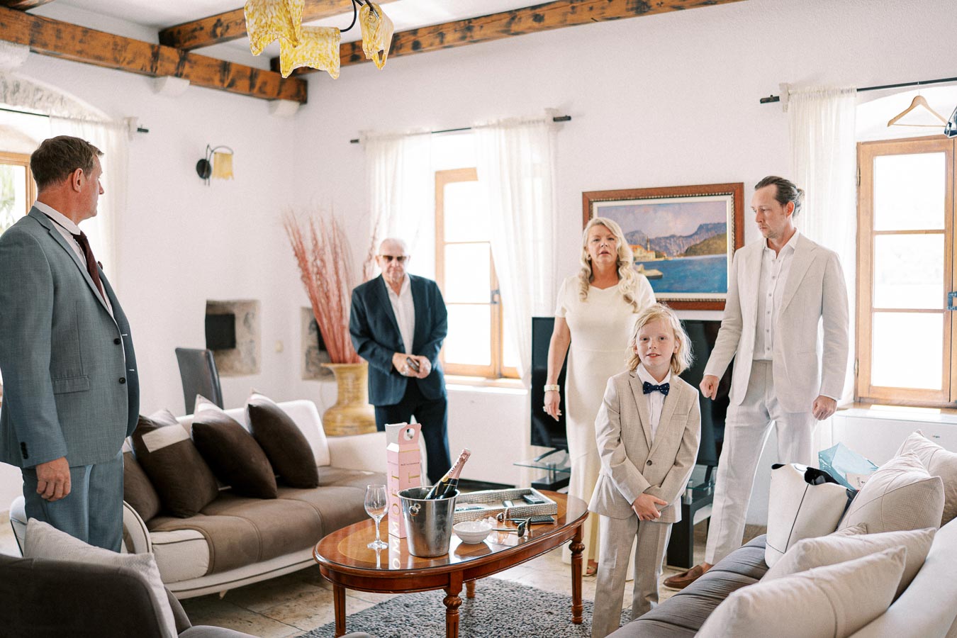 A group of people in elegant attire gather in a stylishly decorated living room with a wooden coffee table, wine and glasses, and natural light streaming through the windows.