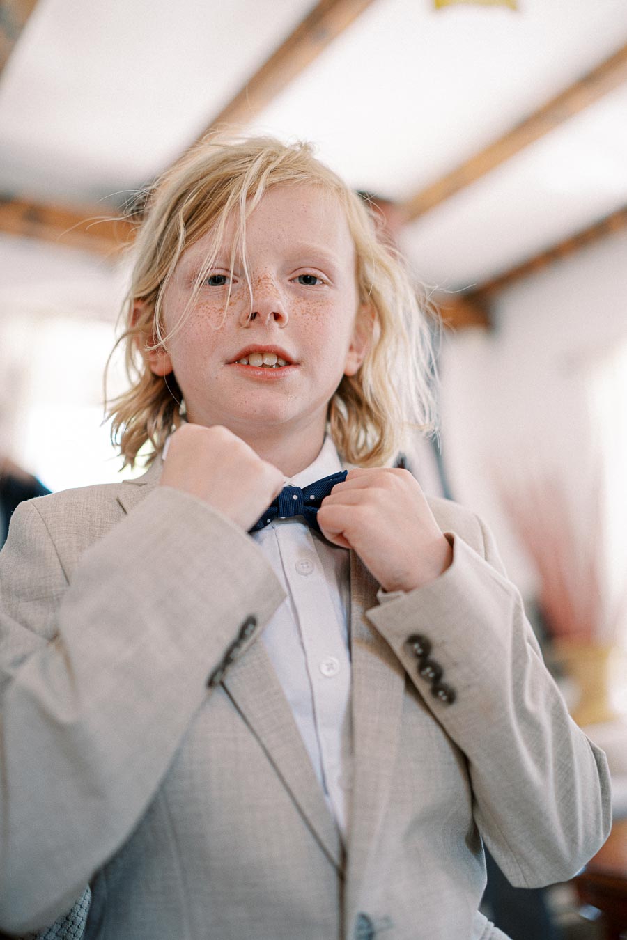 A young child with blond hair confidently adjusts a blue bow tie while wearing a beige suit jacket, smiling slightly in a softly lit indoor setting.