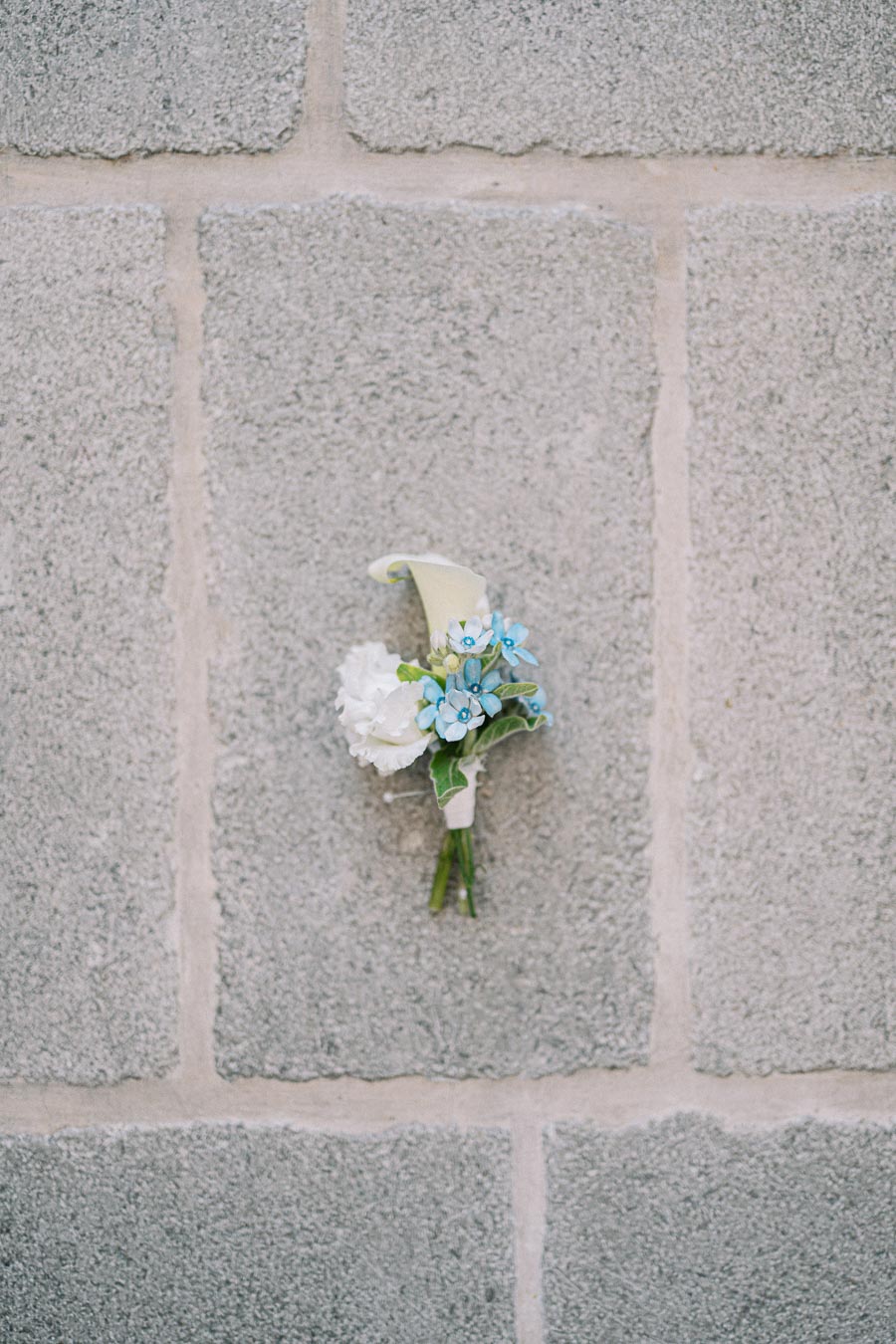 Boutonniere featuring a calla lily, white carnation, and blue flowers with greenery, pinned on a gray brick wall background.