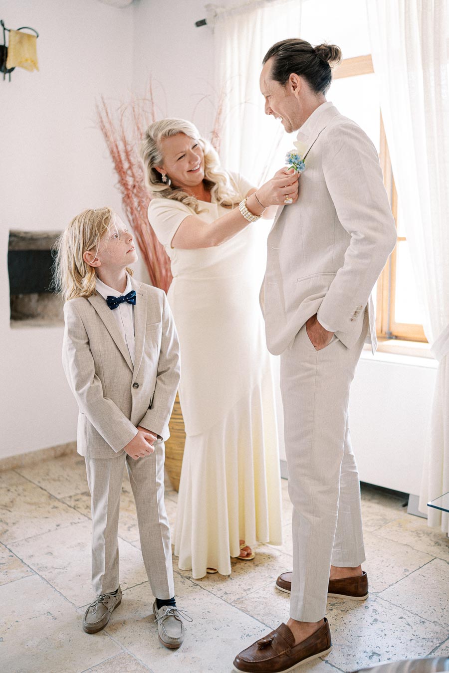 Elegant wedding preparation scene with woman adjusting boutonniere on man's light suit, young boy in bow tie watching, sunlit room with curtains.