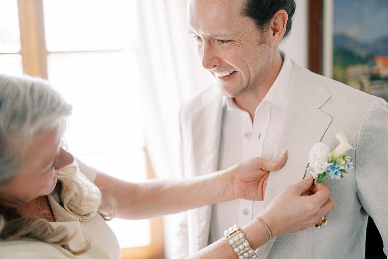 Bride pinning boutonniere on groom's light suit on wedding day, smiling indoors with soft natural lighting.