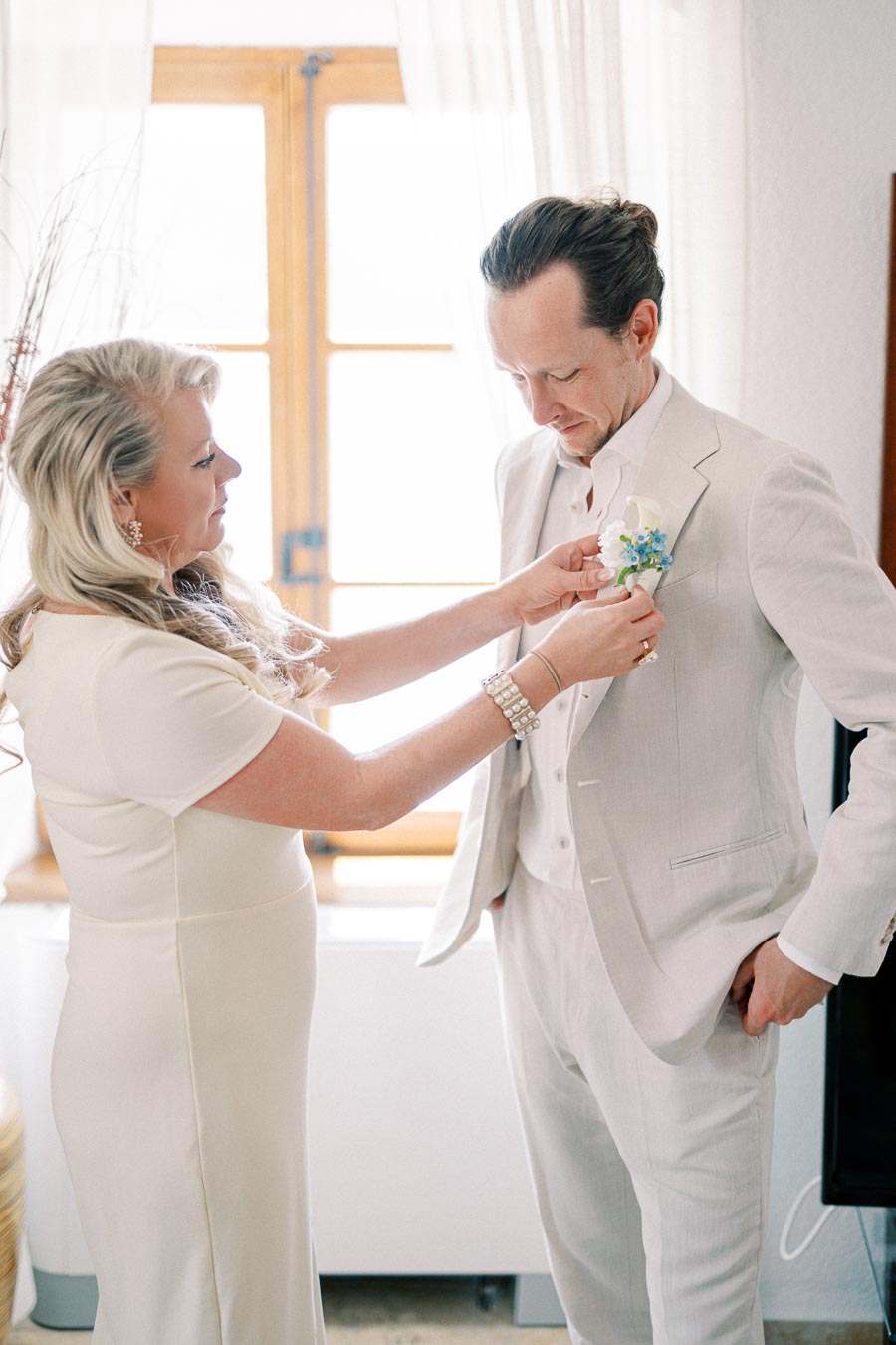 A woman helps a man adjust his boutonniere in a brightly lit room, both dressed in elegant cream attire, suggesting a wedding or formal event preparation.