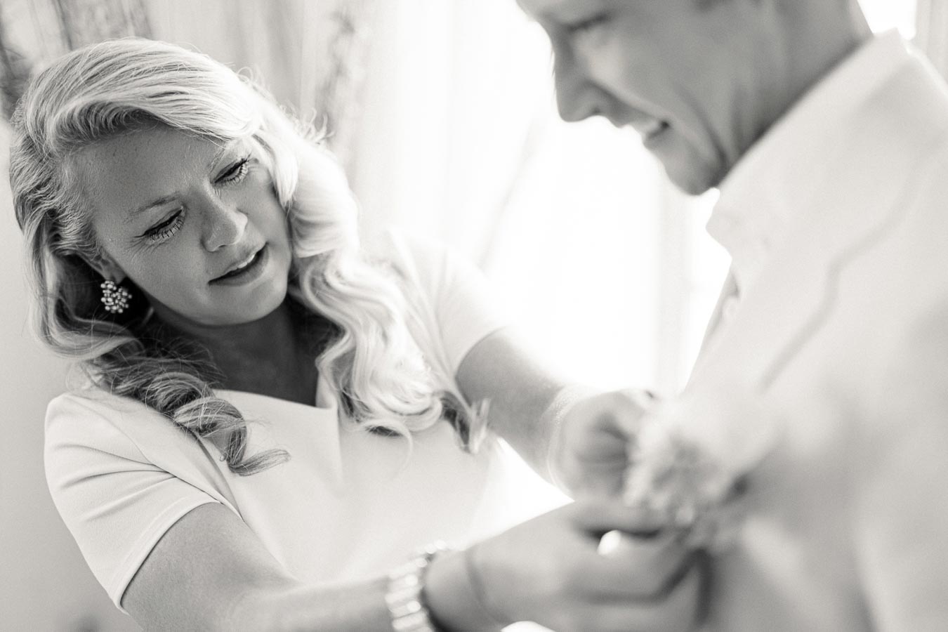 A woman fixing a boutonniere on a man's suit, preparing for a formal event or wedding, in a black and white image.