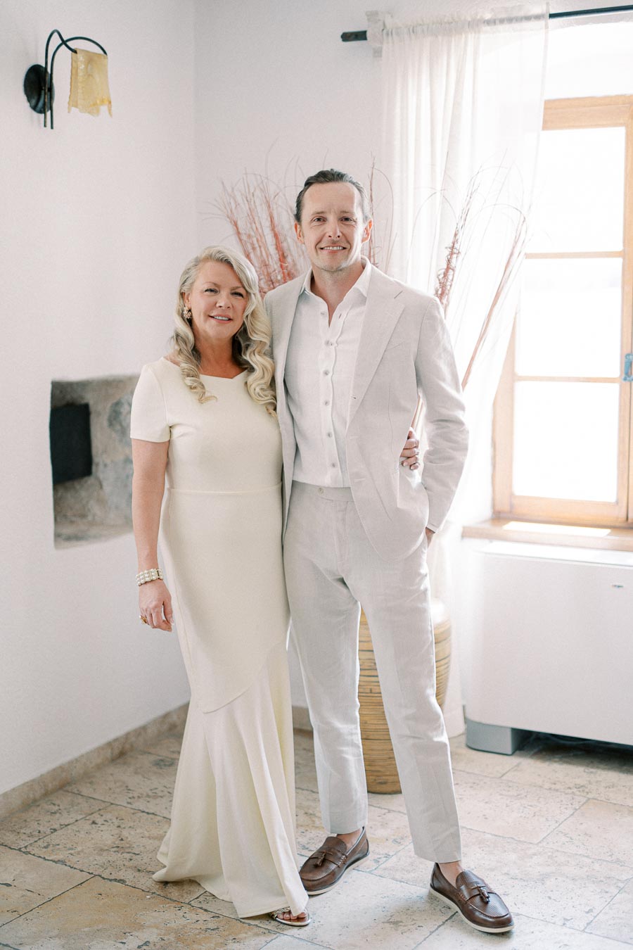 A smiling couple in elegant white attire, standing in a softly lit room with natural light streaming through a window, creating a serene and joyful atmosphere.