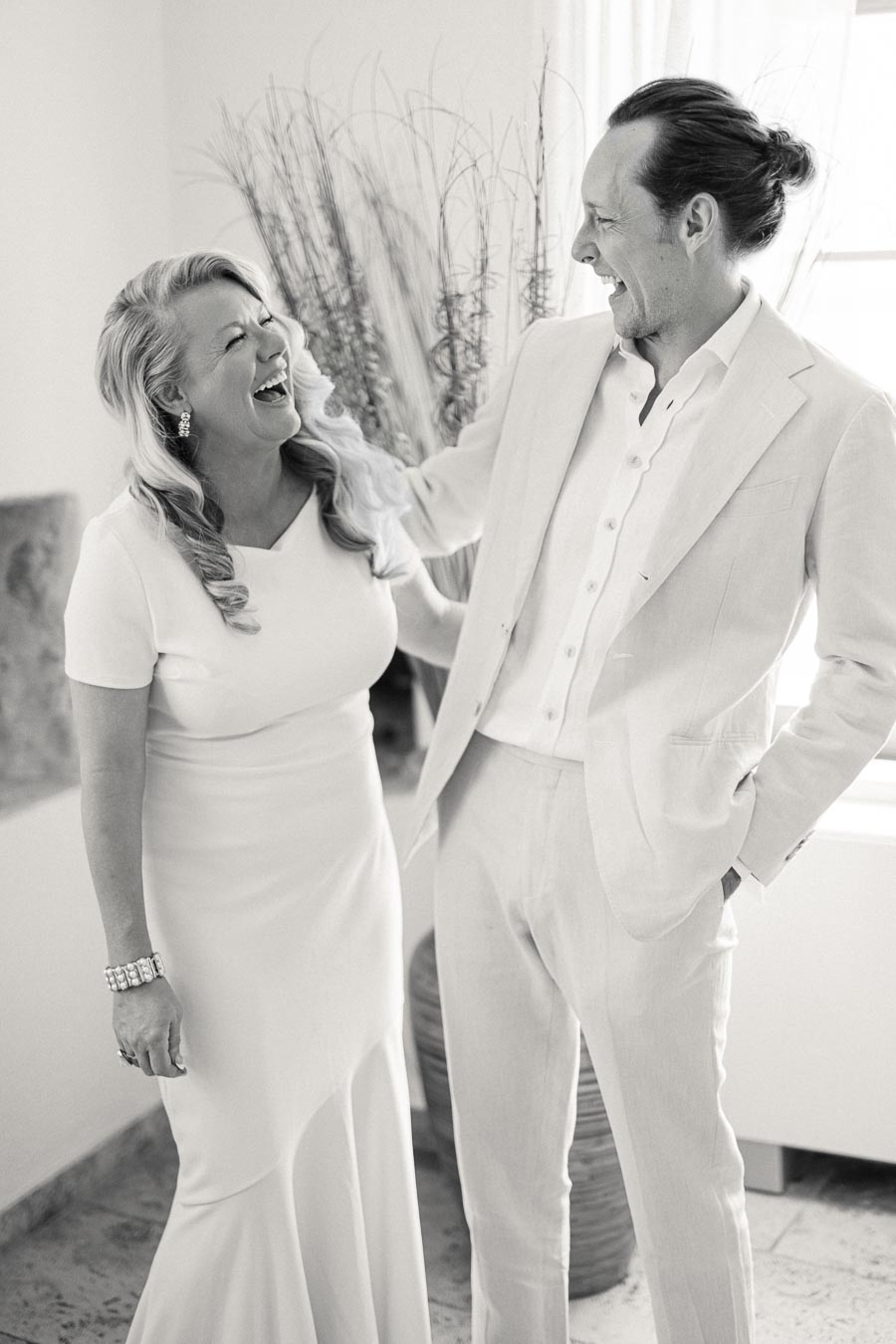 A joyful couple in elegant attire sharing a laugh, dressed in classic, light-colored outfits with natural decor in the background, creating a cheerful and sophisticated ambiance.