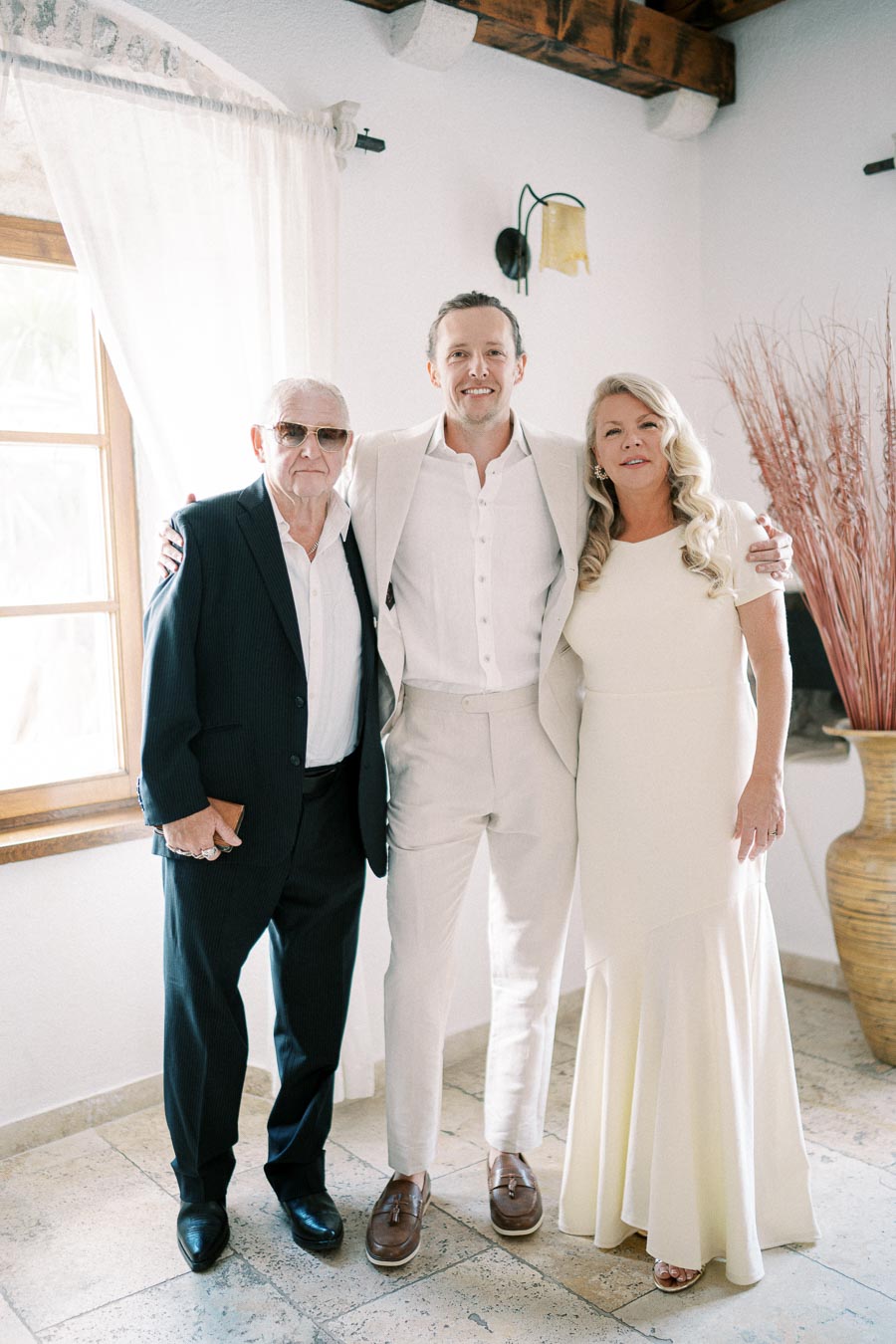 A smiling family group portrait featuring an elderly man in a dark suit, a young man in a white suit, and a woman in a light-colored dress standing in a well-lit room with a tiled floor, a window, and decorative elements in the background.
