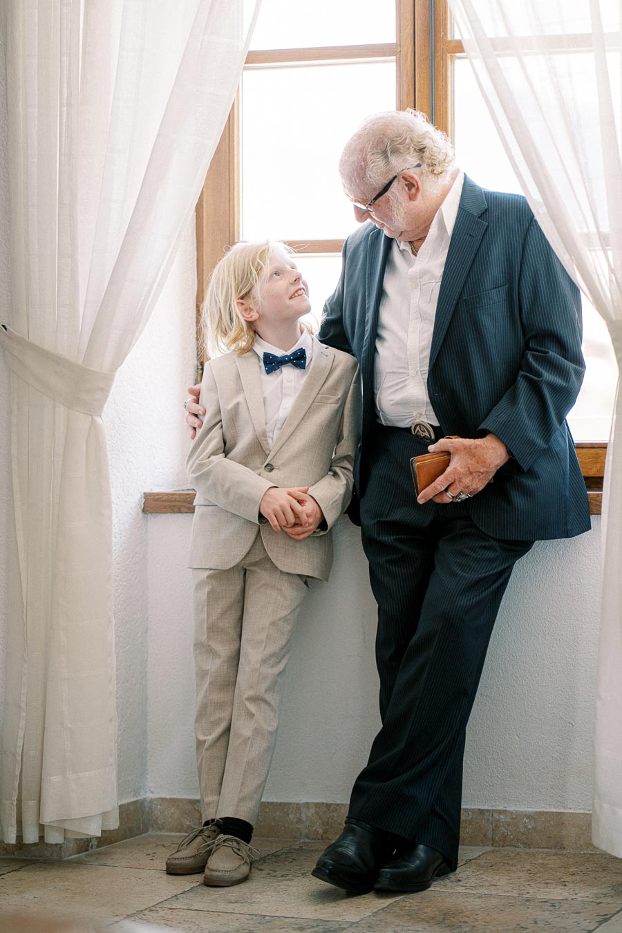 Grandfather and grandson sharing a tender moment in formal attire by a sunlit window, bonding and smiling at each other.