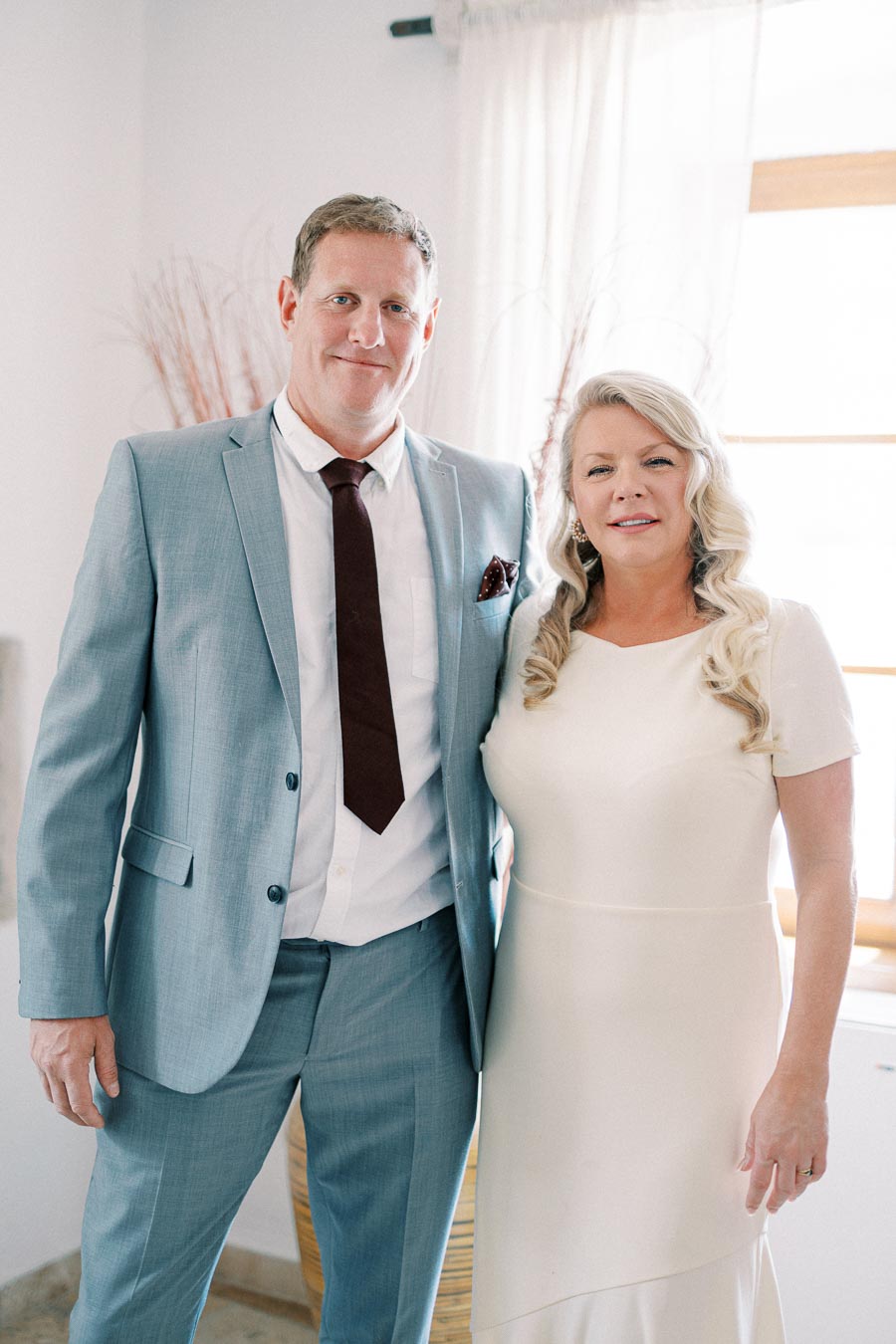 A couple dressed in formal attire, with the man in a light blue suit and the woman in a white dress, standing together in a well-lit room.