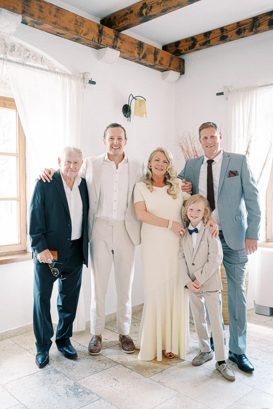 A family dressed in formal attire poses together indoors, featuring a light-colored theme and wooden ceiling beams in the background.