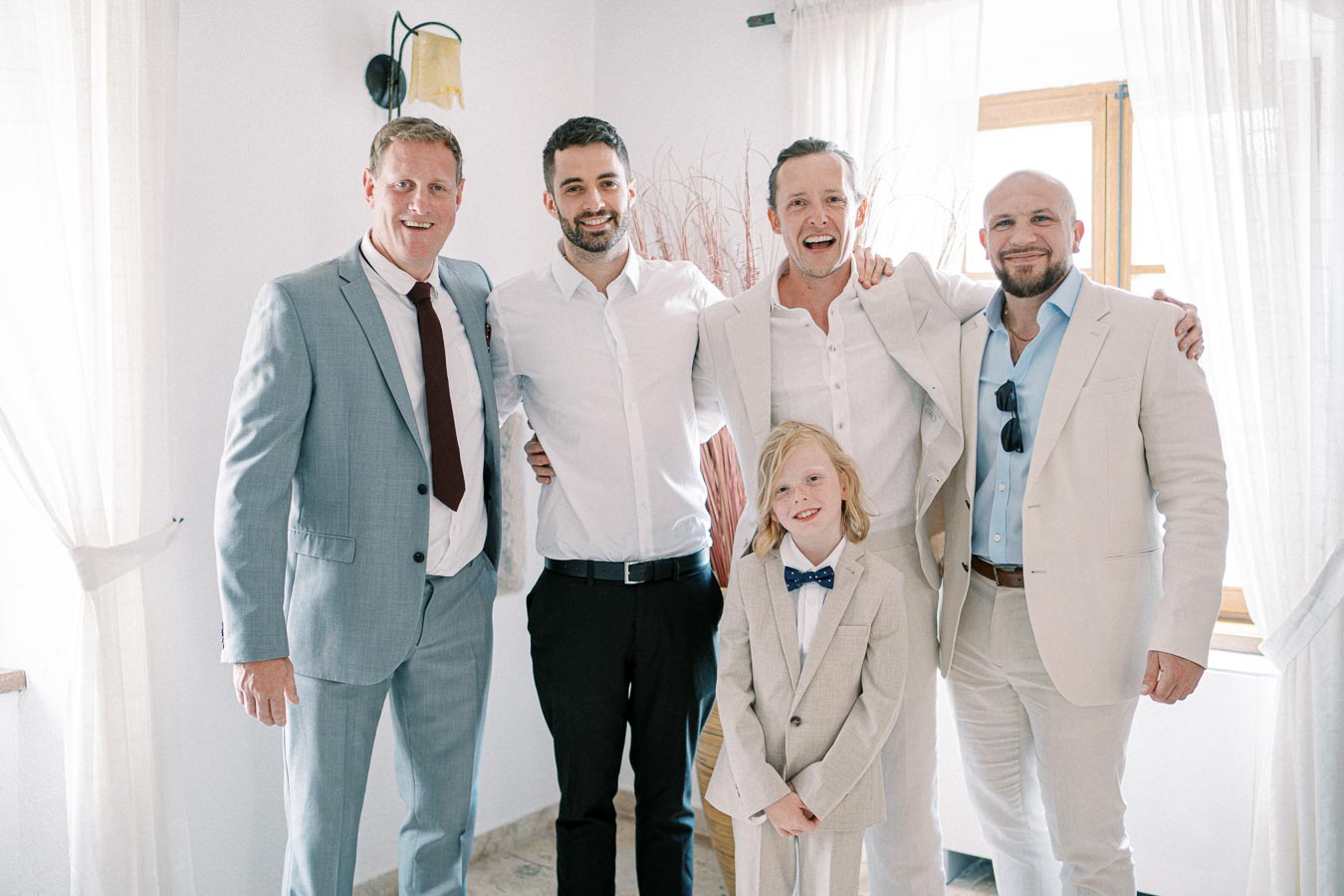 Group of five people dressed in suits and smiling in a bright room with curtains and a window.