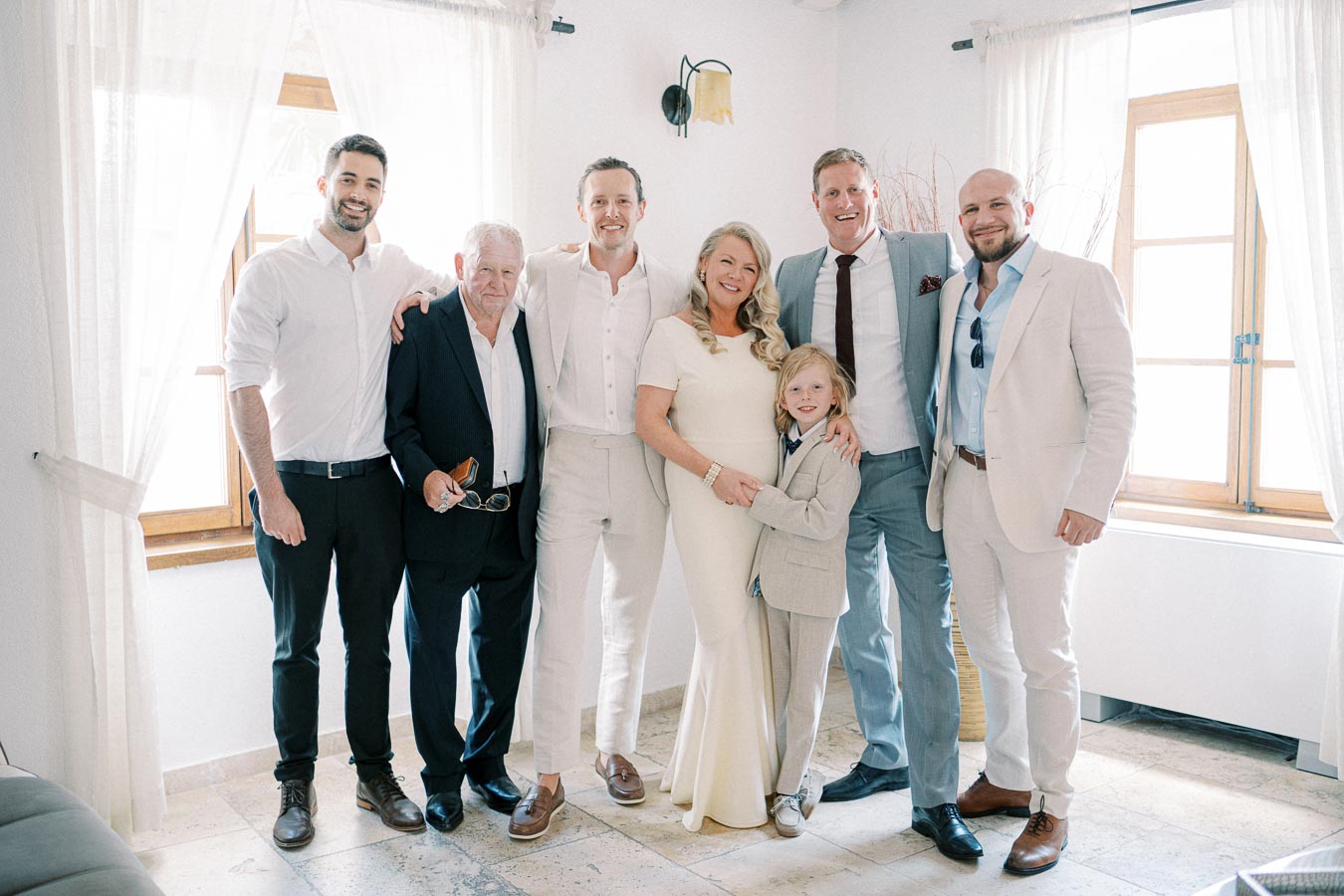 A happy family group poses indoors next to a sunlit window, dressed in formal attire, with a young boy clasping hands with an older woman in a white dress.
