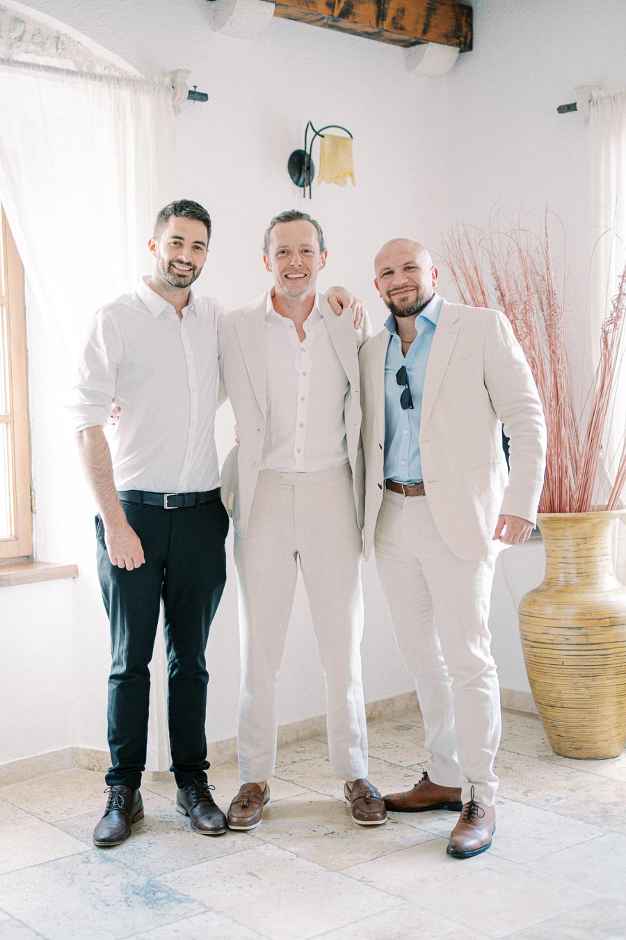 Three men in smart attire standing together, smiling, in a softly lit room with rustic decor and neutral tones.
