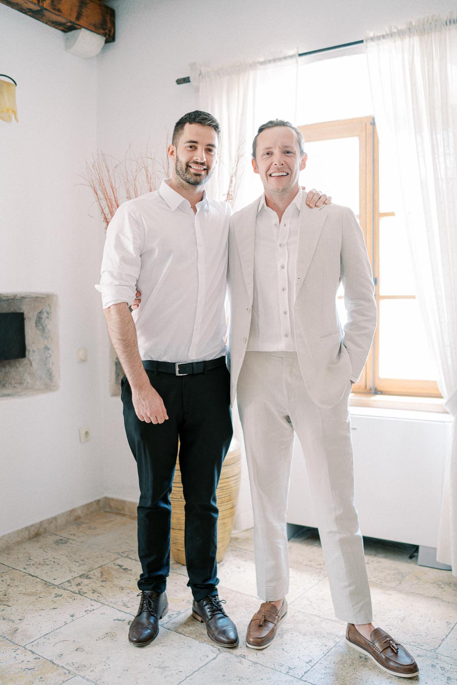 Two men standing indoors, dressed in light-colored formal attire, posing for a photo in a well-lit room with large windows and elegant decor.