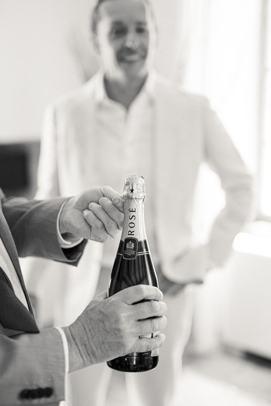 Black and white image of a person opening a bottle of rosé champagne, with another person in a suit blurred in the background, suggesting a celebratory occasion.