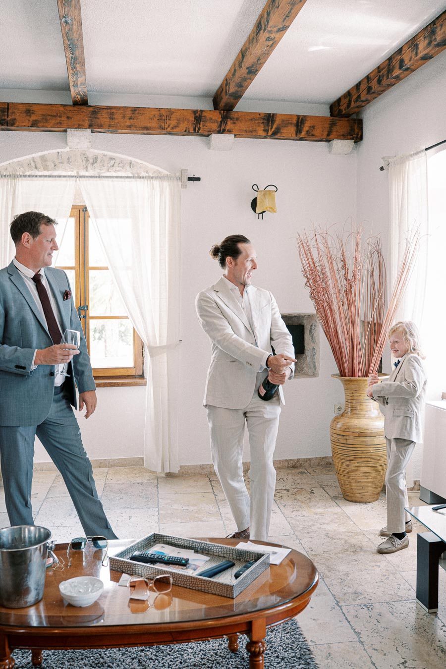 Three people in formal suits celebrating indoors, one holding a champagne bottle, with a stylish room featuring wooden beams and decorative elements.