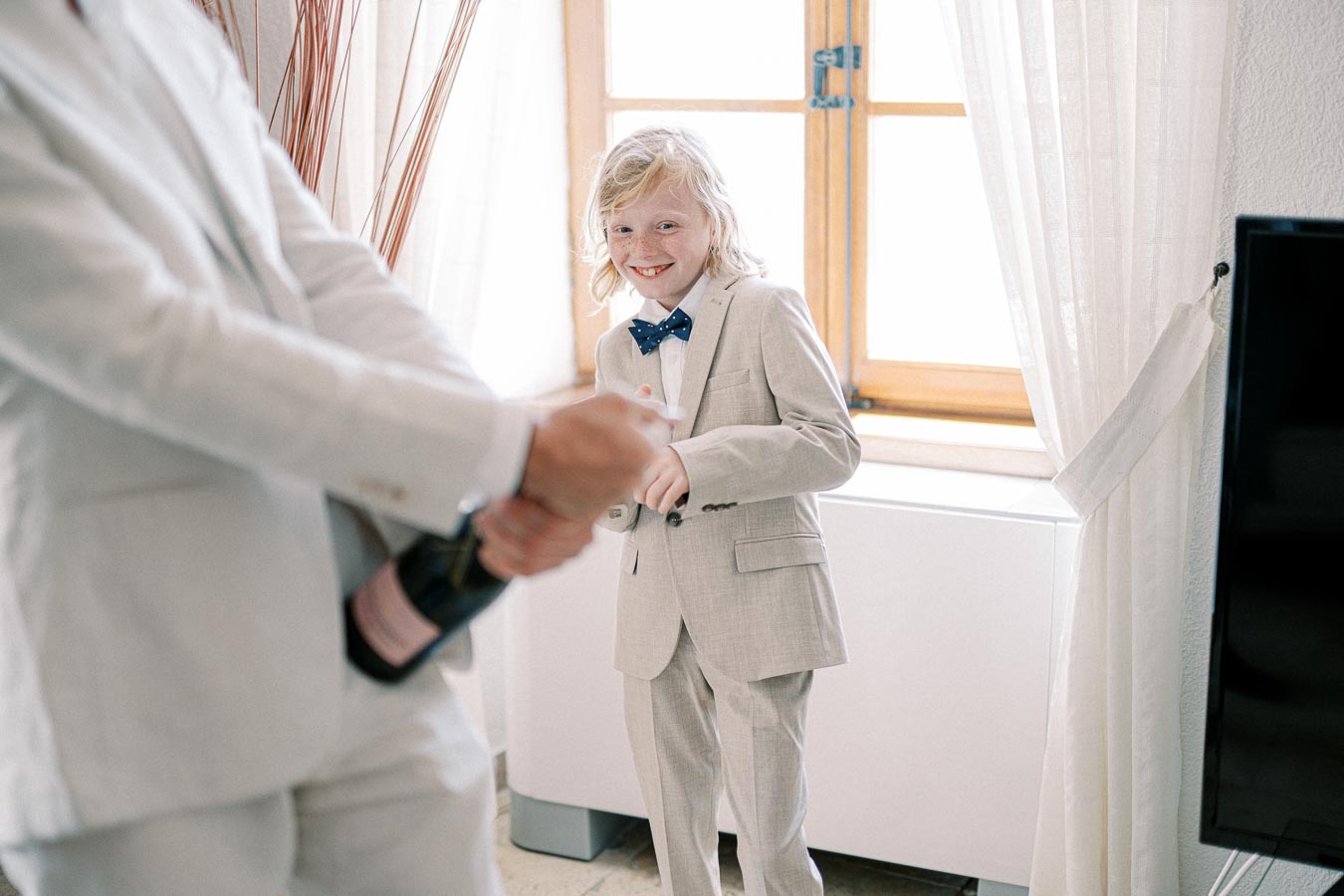A smiling child in a light-colored suit with a blue bow tie stands in a bright room near a window, while an adult in a matching suit is about to open a champagne bottle in the foreground.