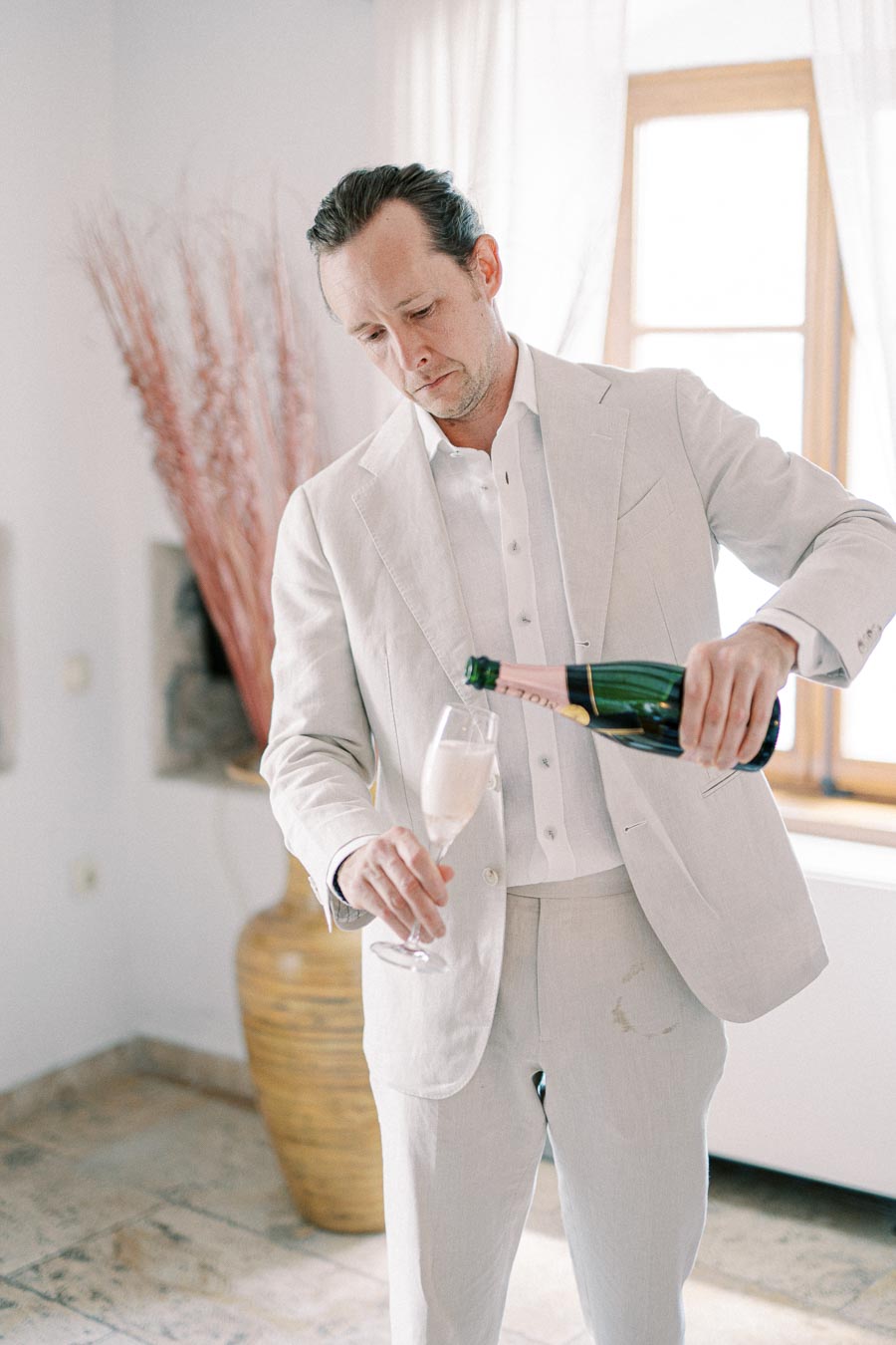 A man in a light-colored suit pours champagne into a flute glass in a bright room with a large window and decorative vase, creating an elegant and sophisticated atmosphere.