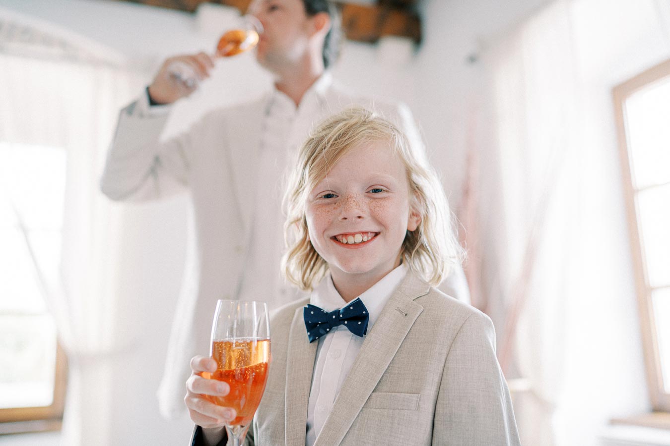 A young boy in a light suit and polka dot bow tie smiles while holding a glass of orange beverage, with a man sipping a drink in the blurred background of a brightly lit room.