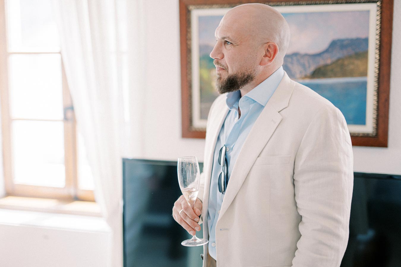 A man in a white blazer holding a champagne glass indoors, looking contemplative with a window and framed art in the background.