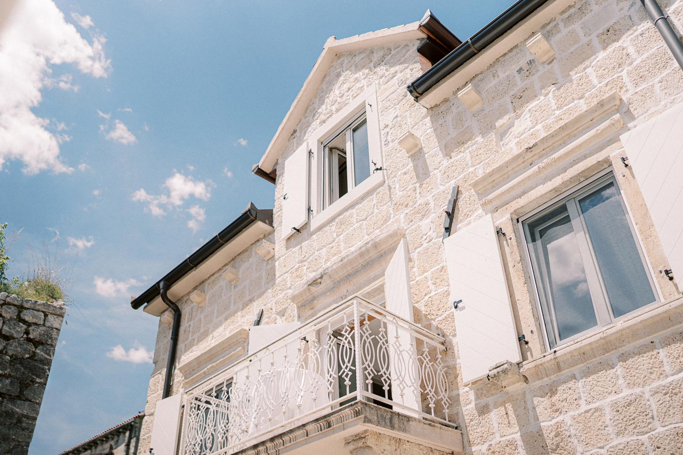 Elegant stone house facade with white shutters and ornate balcony railing under a clear blue sky.