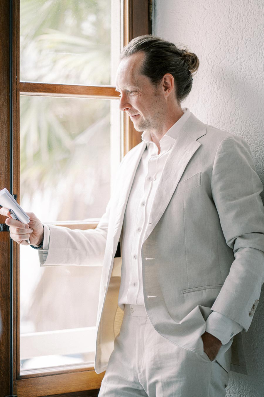 Man in a light suit reading a book by a sunlit window, exuding a relaxed and thoughtful demeanor.