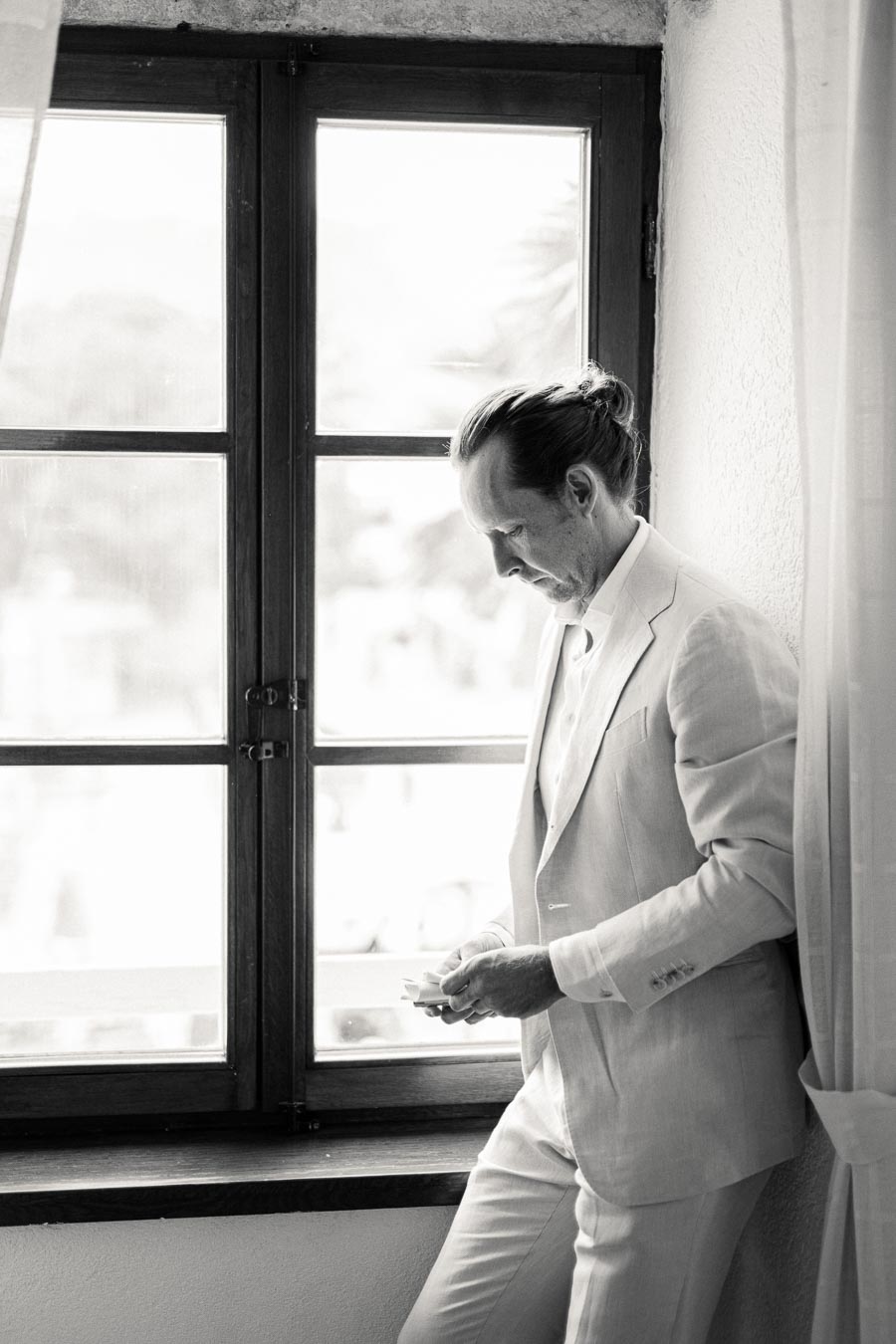 Black and white photo of a man in a light-colored suit standing by a window, looking down thoughtfully while holding a phone.