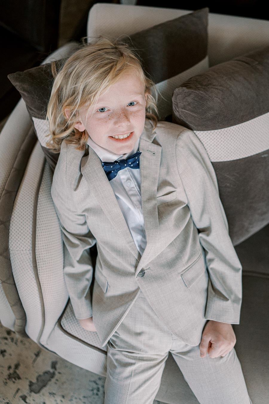 Young child in a light gray suit and blue bowtie sitting on a sofa, smiling confidently at the camera.