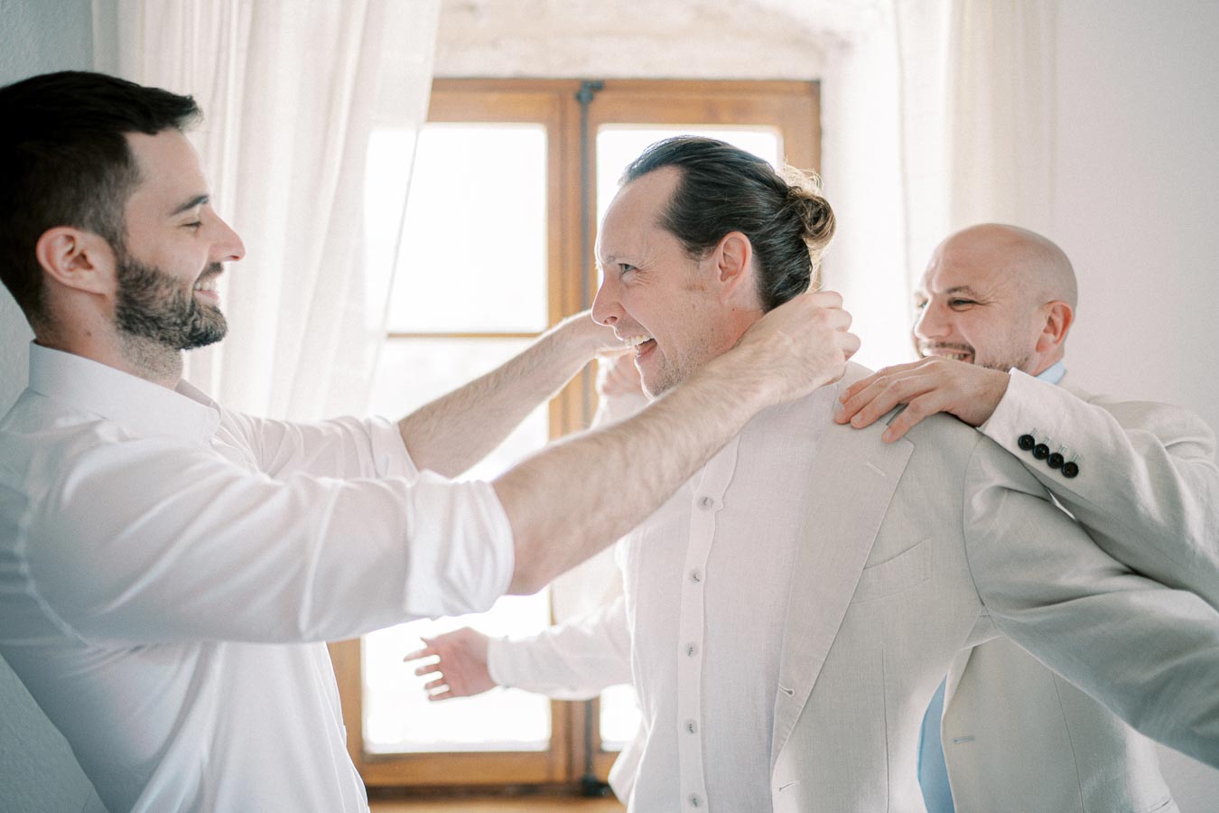 Three men in a light-filled room, joyfully helping one man with a suit jacket, suggesting a wedding preparation or a formal event.