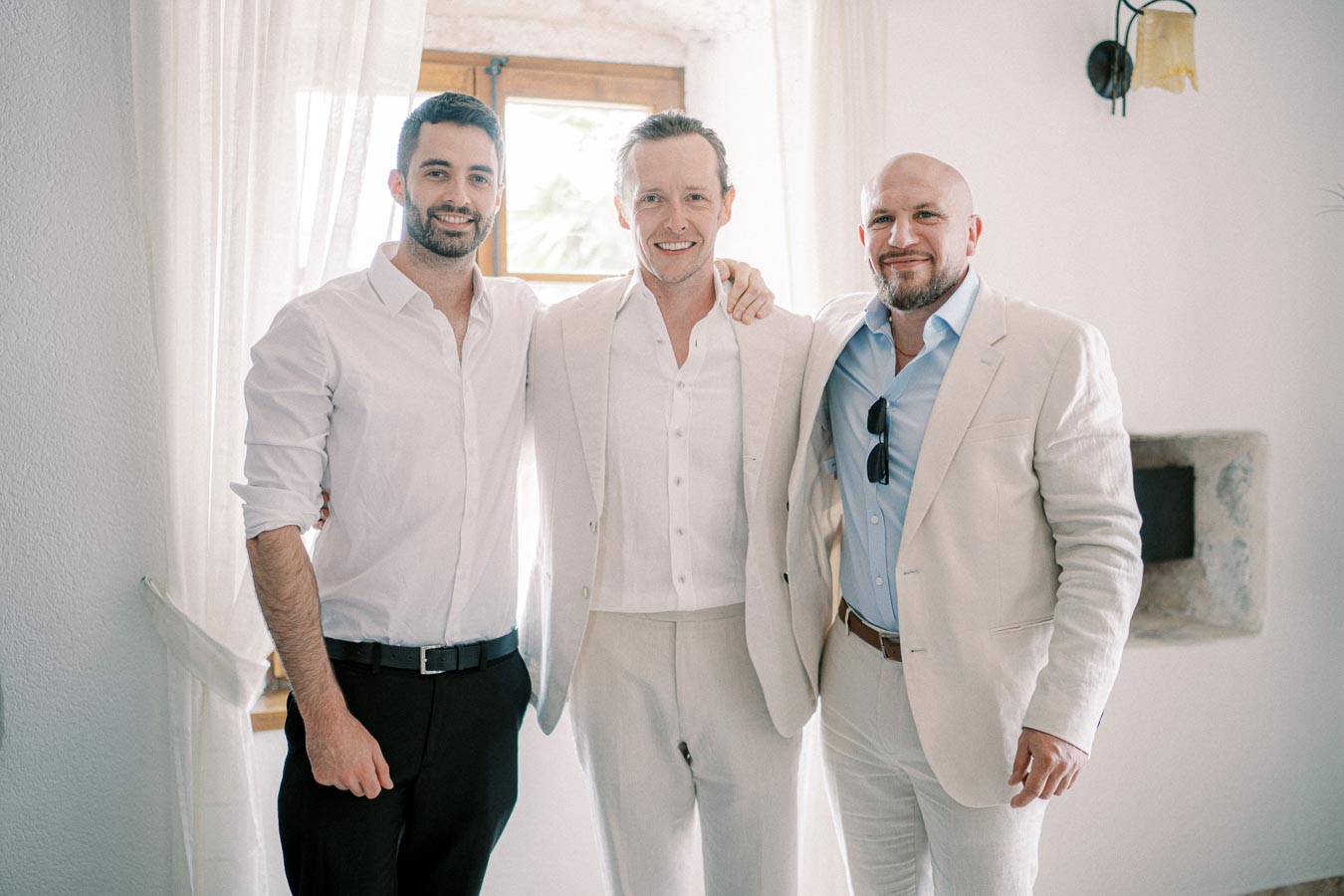 Three men dressed in light-colored formal suits, smiling and posing together indoors near a window with sheer curtains.
