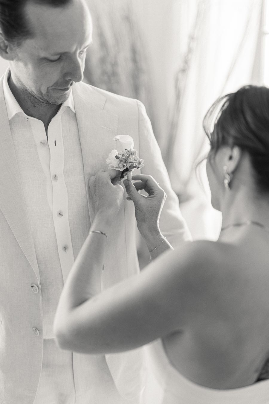 A woman adjusts a boutonniere on a man's light-colored suit jacket, captured in elegant black and white photography.