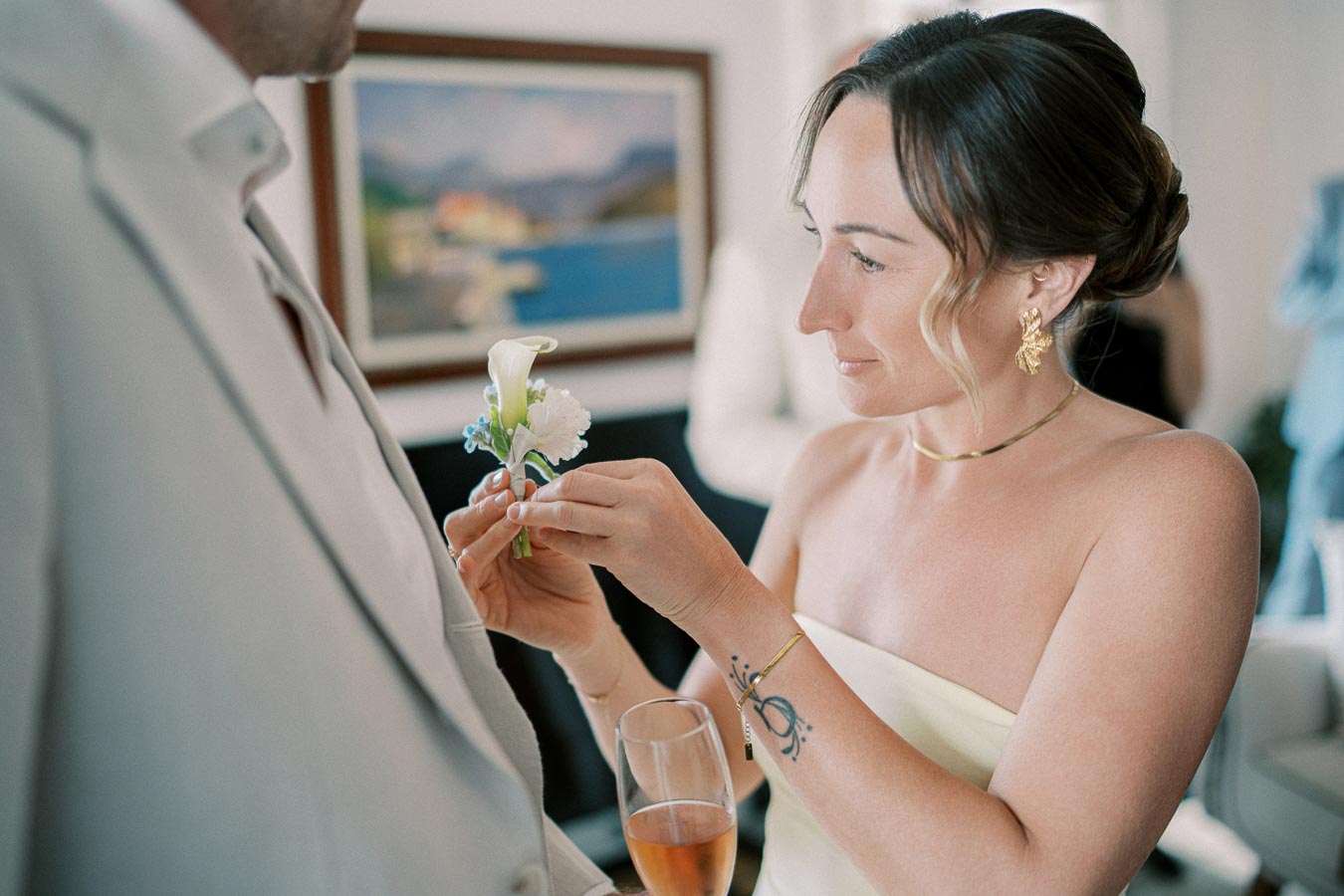 Woman attaching boutonniere to man in light suit at an elegant event, holding a champagne glass.