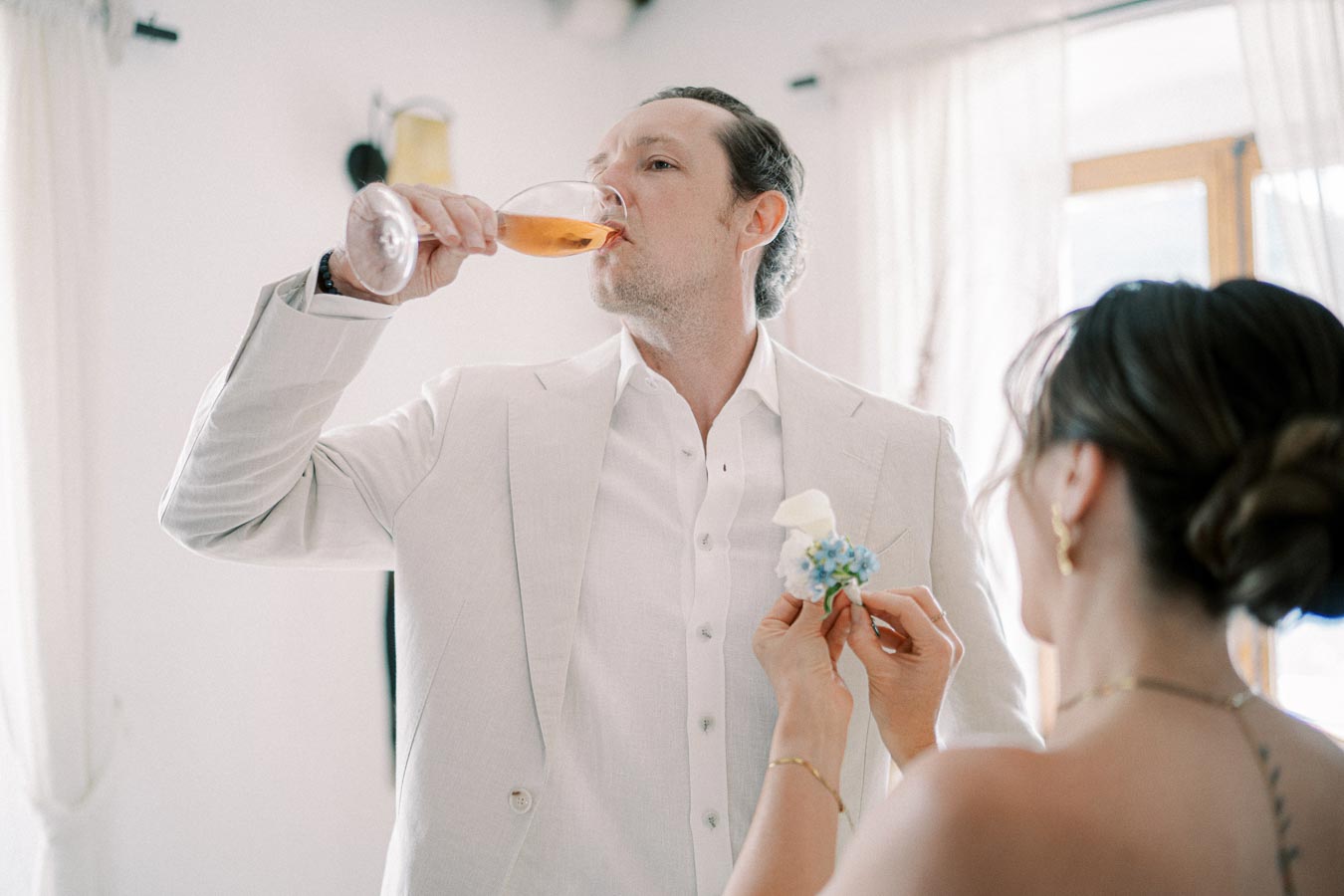 Man in white suit drinking rosé wine at a wedding while a woman pins a boutonniere on him.