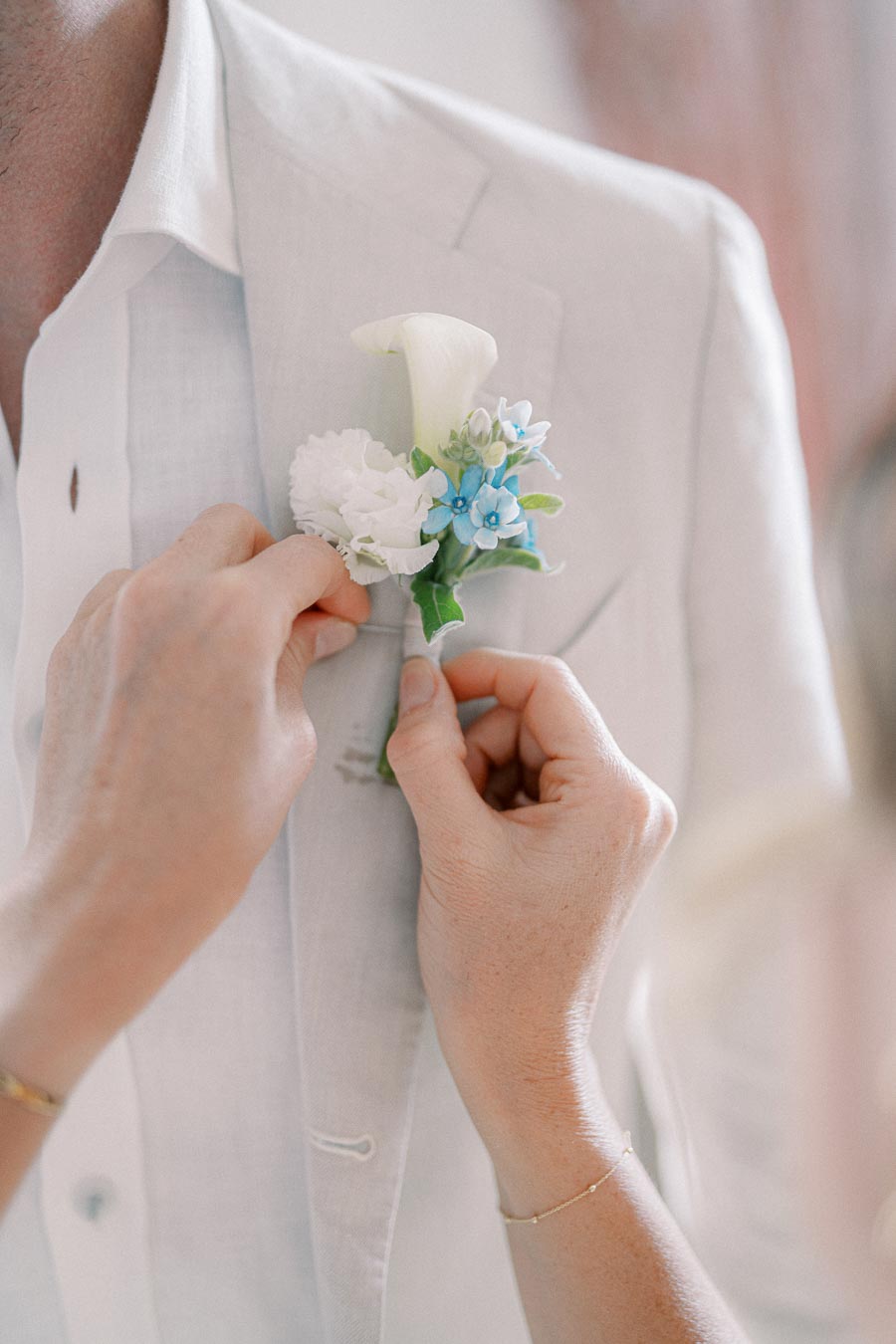 Close-up of a person pinning a boutonniere with white and blue flowers onto a light gray suit jacket.