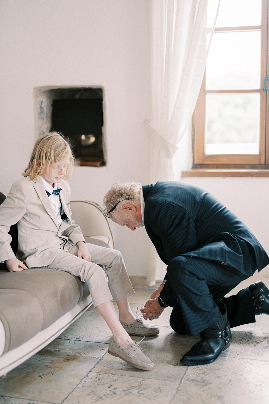 Elderly man helping young boy in a suit tie his shoelaces in a living room, creating a warm, family bonding moment.