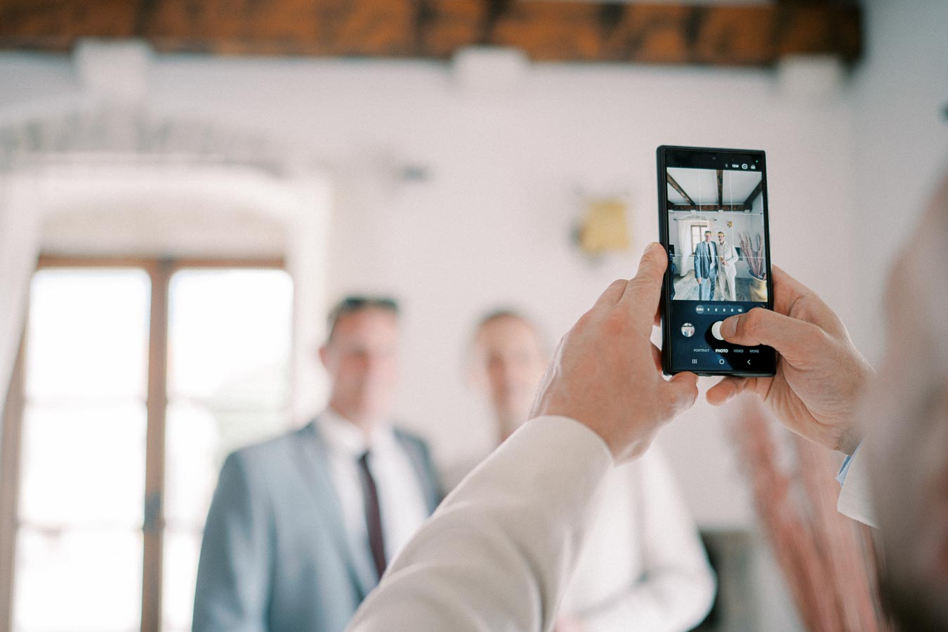 A person taking a photo with a smartphone of two men dressed in suits, standing indoors in a well-lit room with large windows.