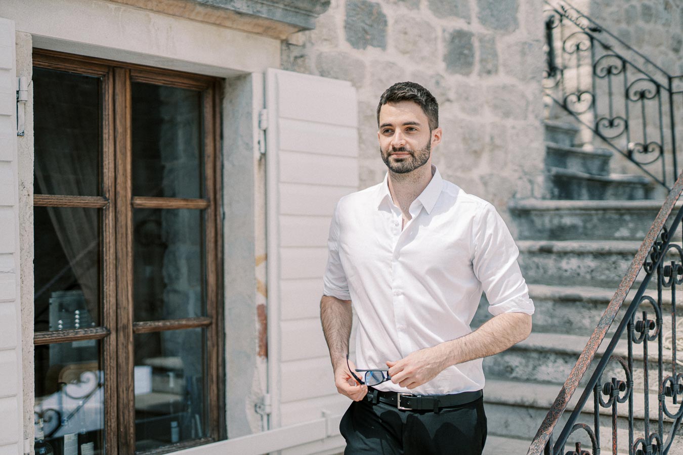 A man in a white shirt holding glasses, standing next to a rustic stone building with a wooden window and stairs in the background.