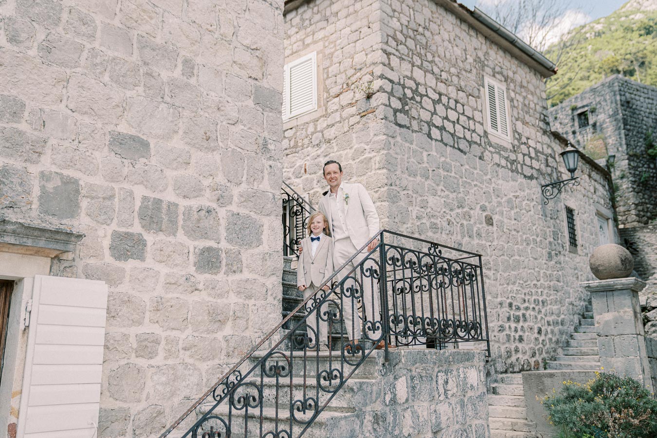 Father and son in matching beige suits standing on stone staircase of historic stone building.