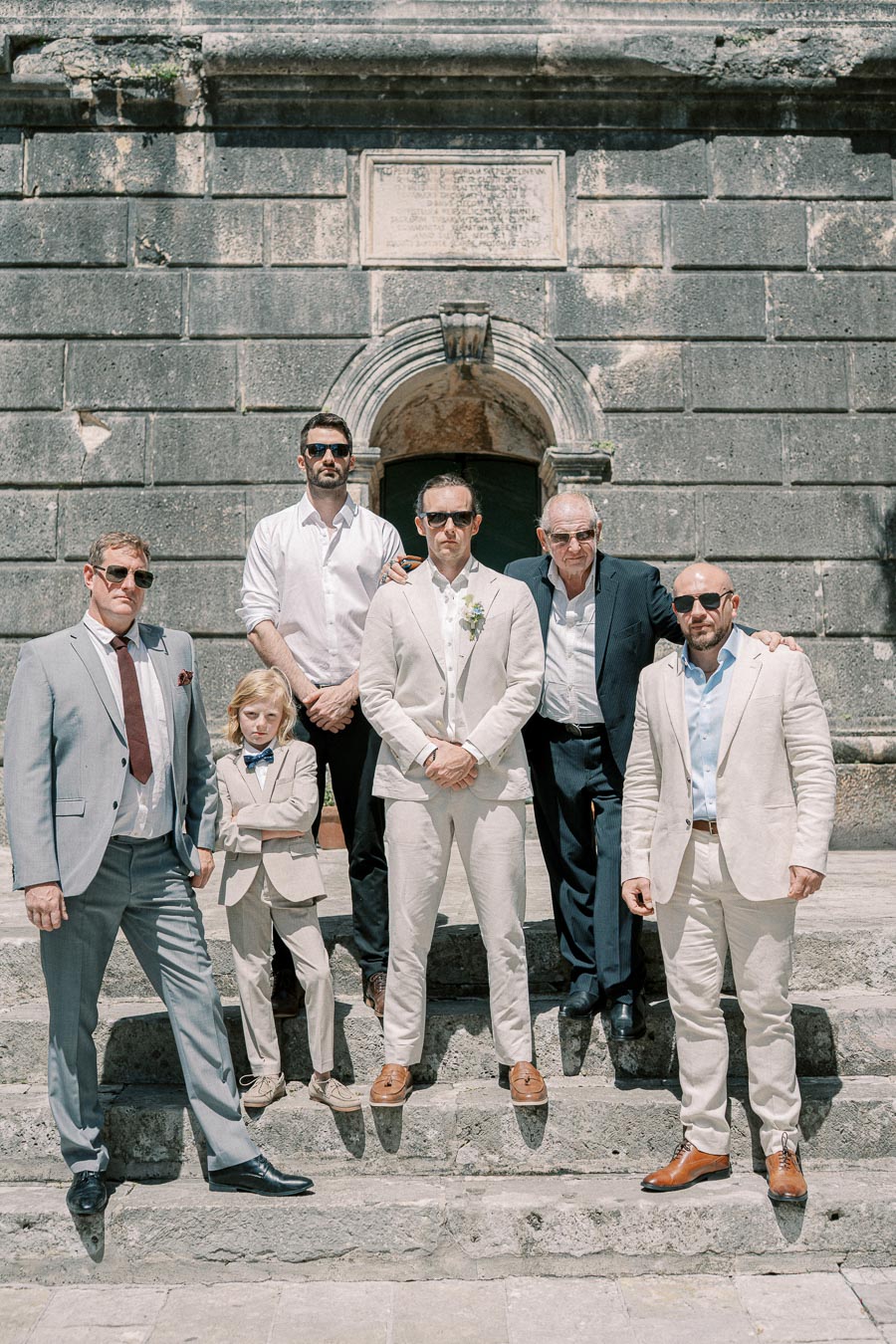 Group of men and one child wearing suits and sunglasses, posing on stone steps in front of an old stone building, conveying a stylish and formal atmosphere under sunny weather.