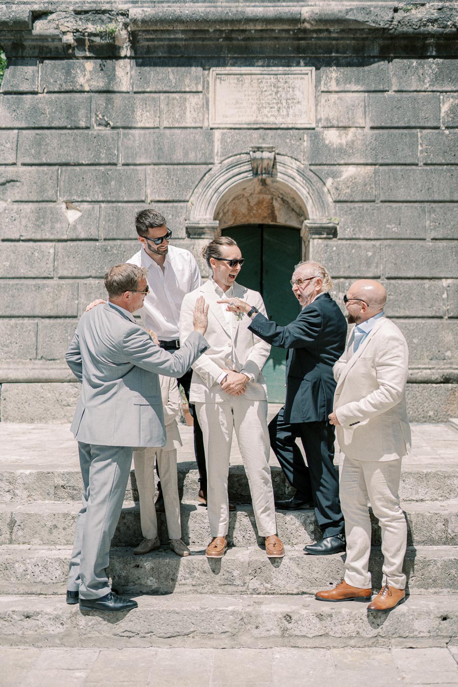 A group of well-dressed men in suits, gathered together and chatting on stone steps in front of a historic building.