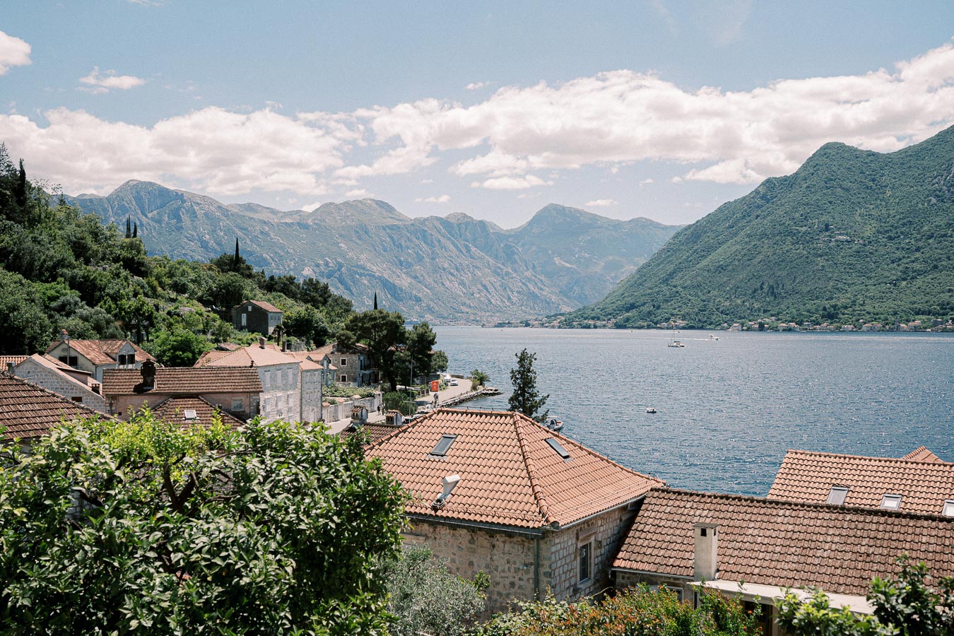 Scenic view of Kotor Bay in Montenegro, featuring rustic terracotta rooftops, lush green hills, and majestic mountains under a clear blue sky.