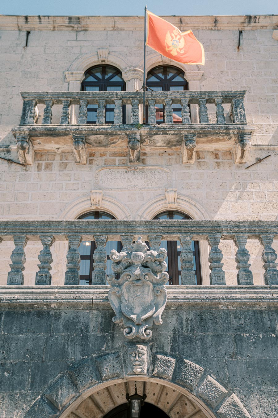 Historic stone building facade with arched windows and ornate balcony, featuring a national flag atop and intricate coat of arms engraving, set against a clear blue sky.
