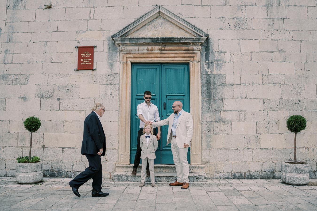 A group of people dressed in formal attire, including a young boy in a suit, standing in front of an old stone building with a teal door in a historic town square.