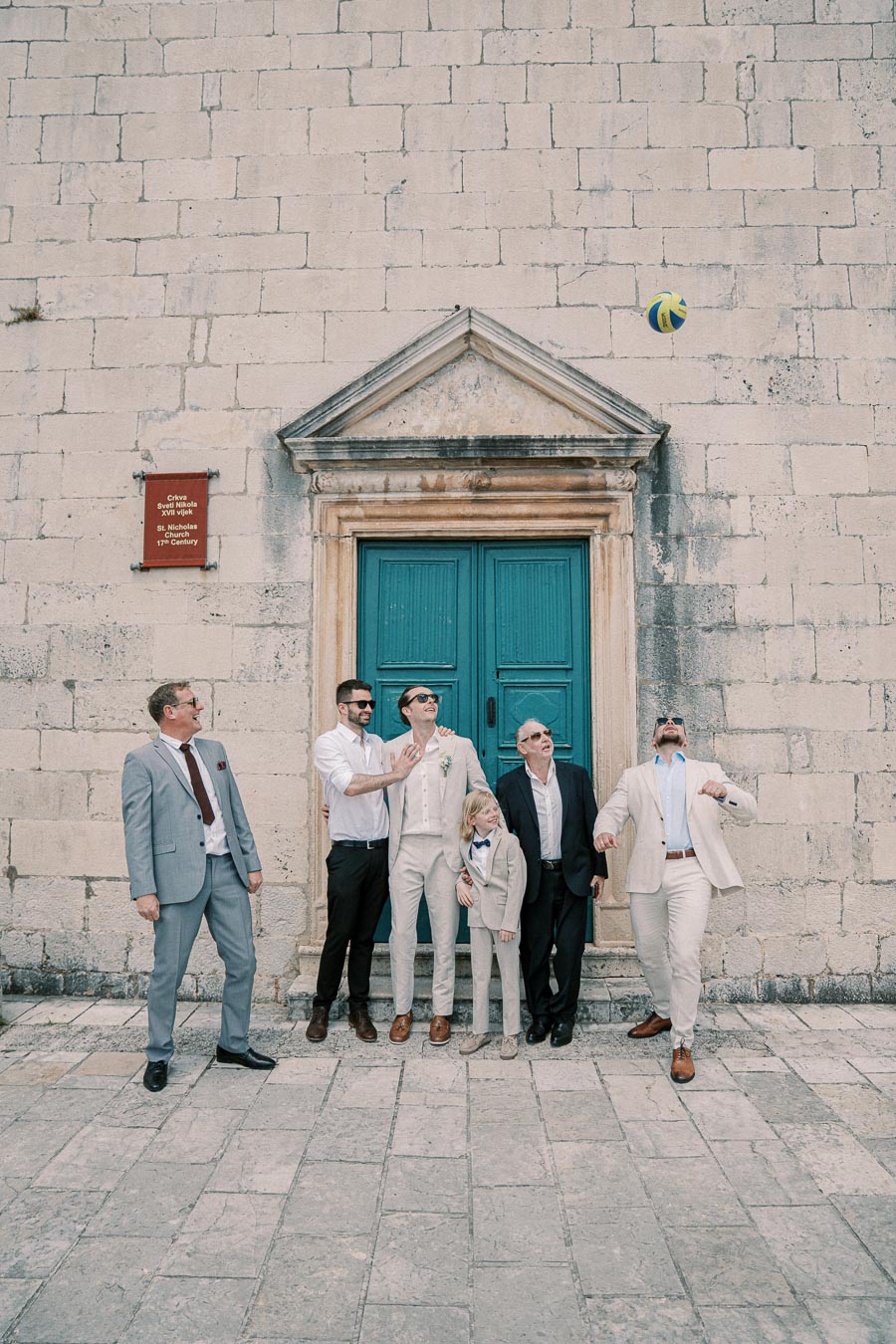 Group of men and a child dressed in formal attire, posing playfully with a ball outside a historic stone building with a teal door.