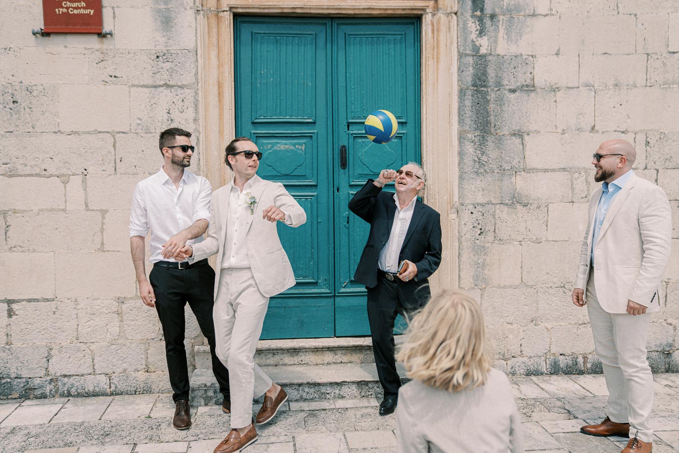 A group of men dressed in semi-formal attire playfully interacts in front of a historical stone building with a vibrant blue door. One man is about to catch a blue and yellow volleyball, while others watch and laugh. The setting suggests a relaxed, joyful outdoor event with a hint of elegance.