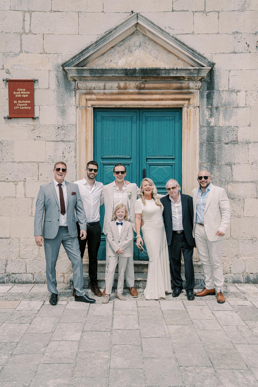 A group of elegantly dressed people posing in front of a historic building with a turquoise door, likely at a wedding gathering.