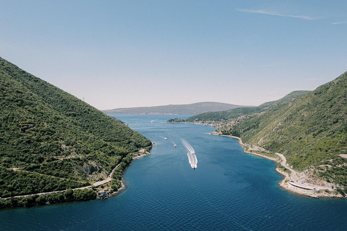 Aerial view of a picturesque blue bay with boats creating white trails on the water, flanked by lush green hills and a winding coastal road under a clear sky.