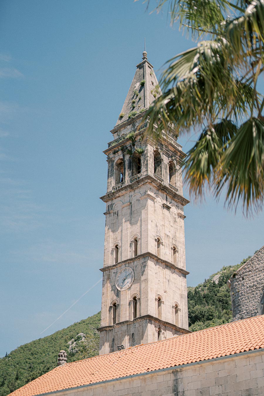 Historic stone bell tower with clock against a clear blue sky in a scenic Mediterranean setting, framed by palm tree leaves.