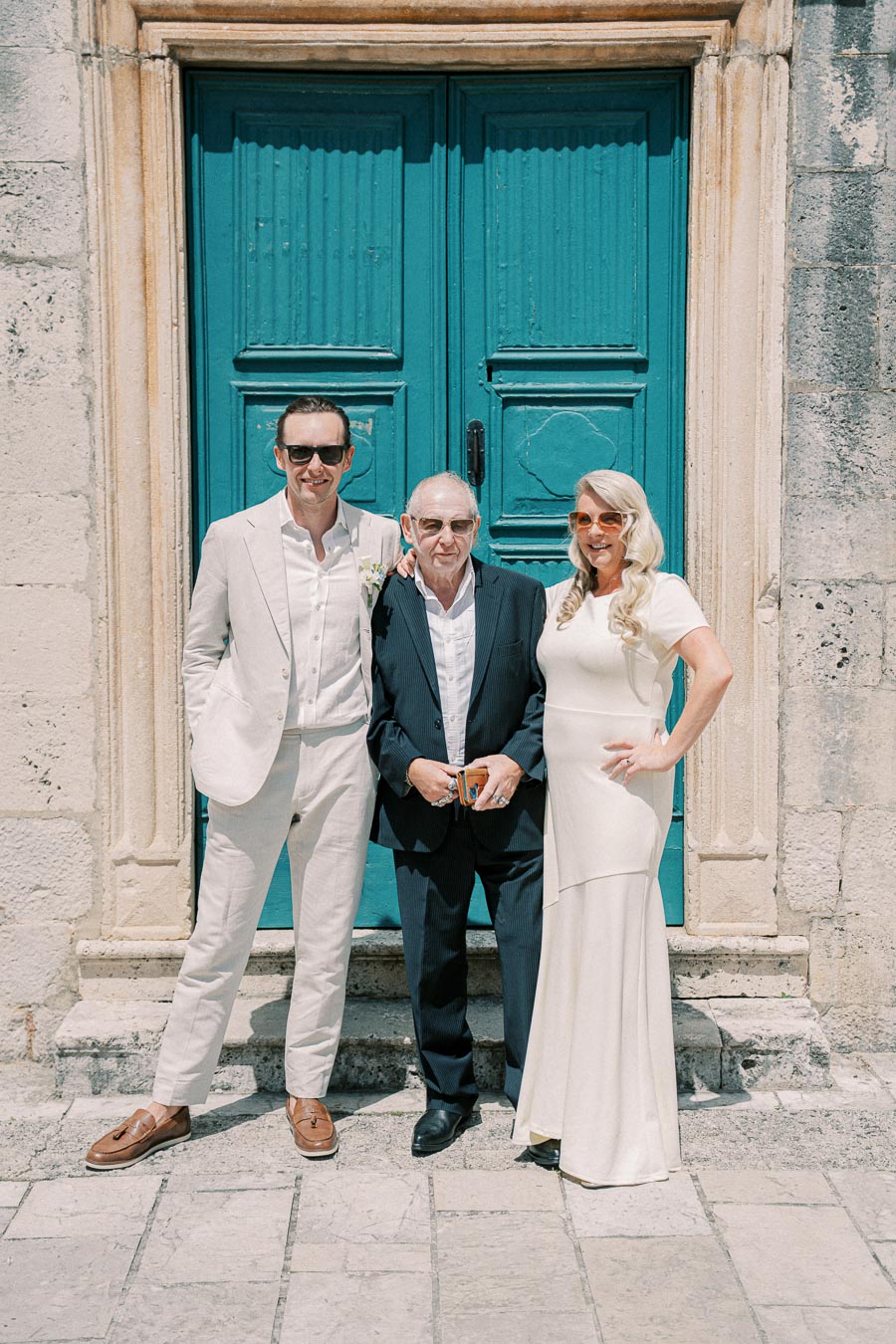 Three people posing in front of a teal wooden door. The man on the left is dressed in a light suit, the man in the center is wearing a dark suit, and the woman on the right is in a white dress. The setting appears to be outdoors with a stone wall backdrop.