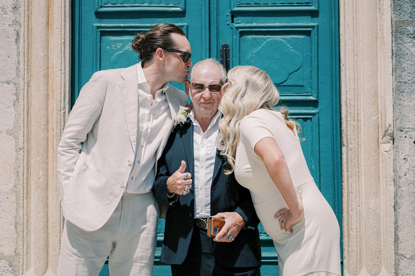 Three people standing in front of a turquoise door; a young man in a cream suit and sunglasses kisses an older man in the center on the cheek, while a woman in a white dress does the same on the other side, conveying a sense of family celebration and affection.