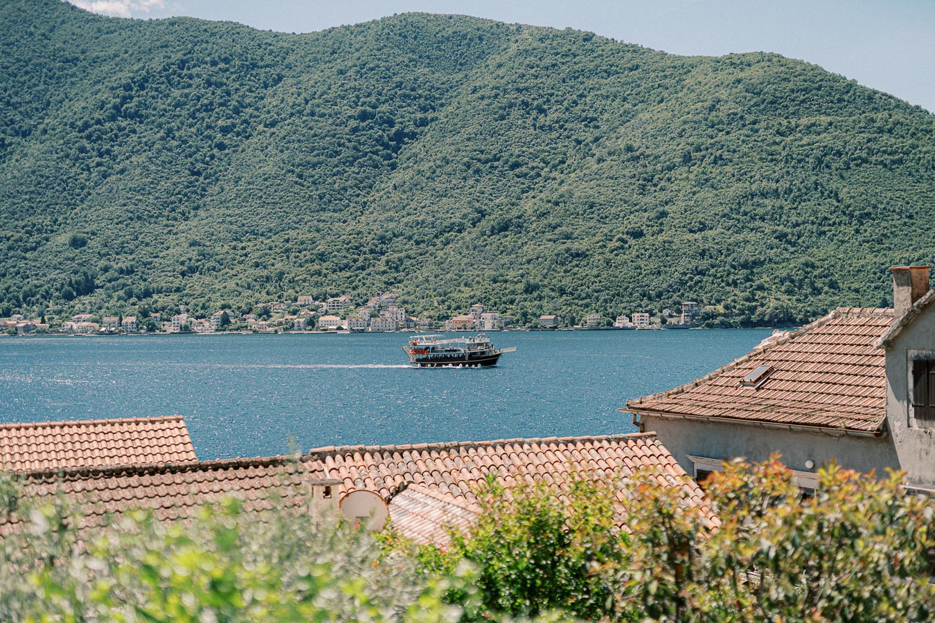 Serene coastal landscape with a boat sailing on a blue sea, surrounded by lush green hills and traditional terracotta-roofed buildings in the foreground.
